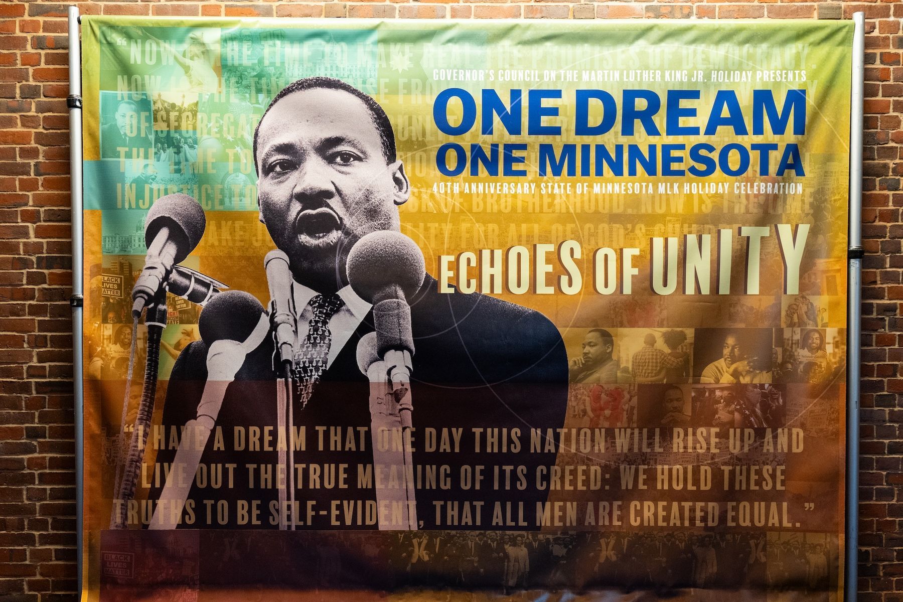 Banner featuring a black and white image of Martin Luther King Jr. speaking into multiple microphones. Text reads: "Governor's Council on the Martin Luther King Jr. Holiday presents One Dream One Minnesota, 40th Anniversary State of Minnesota MLK Holiday Celebration, Echoes of Unity." Background shows faded images of civil rights marches and people, along with the quote: "I have a dream that one day this nation will rise up and live out the true meaning of its creed; we hold these truths to be self-evident, that all men are created equal."